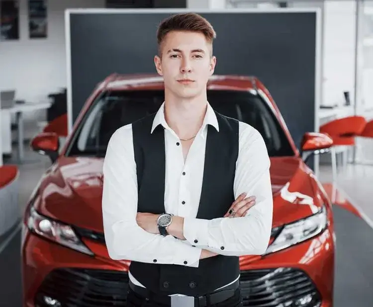 Ein Junger Mann, mit verschränkten Armen, der an einem roten Auto lehnt und selbstbewusst in die Kamera blickt. Autohändler Gießen.